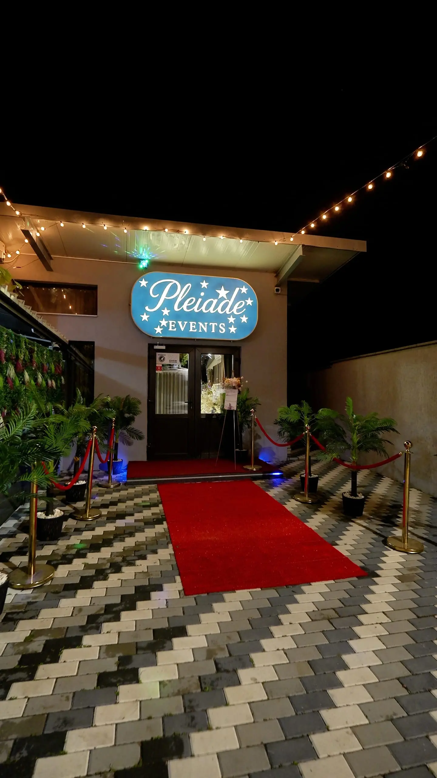 Intrarea în salon, seara, cu un covor roșu până la ușă, stâlpi aurii cu funii roșii și palmieri în ghivece. Deasupra ușii, firma luminoasă Pleiade Events, iar de-a lungul acoperișului, o ghirlandă de becuri.