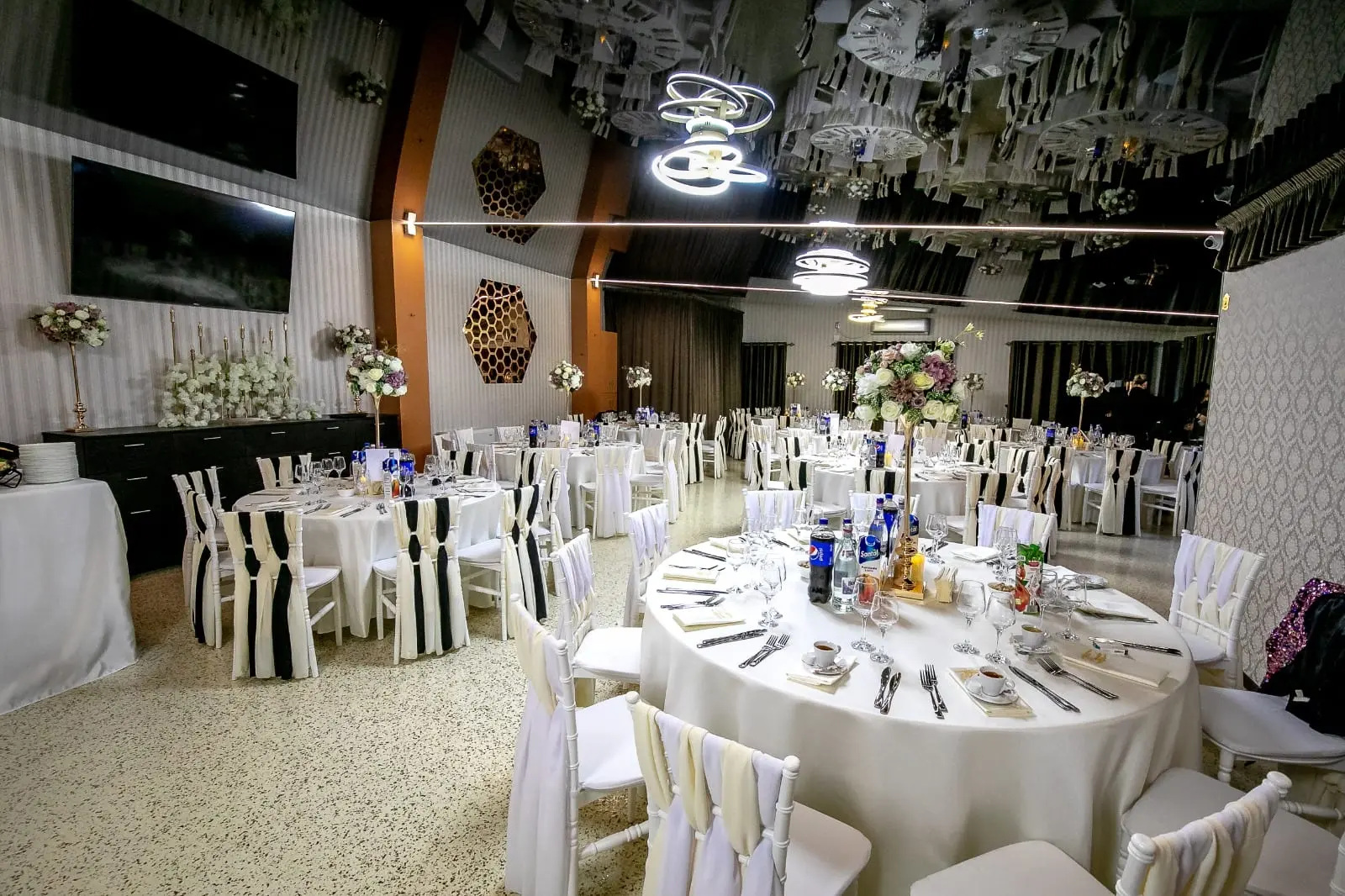 Masă rotundă aranjată, cu aranjament floral înalt pe suport auriu, în sala luminată de lustre LED circulare. Pe perete, o oglindă hexagonală.