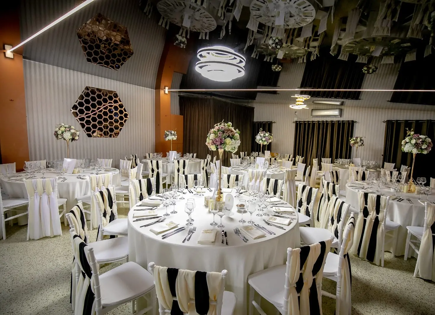Sala cu mese rotunde și scaune albe cu eșarfe crem și negre, sub lustre LED circulare aprinse. Pe perete este o oglindă hexagonală, iar tavanul negru lucios reflectă mesele și oglinda.