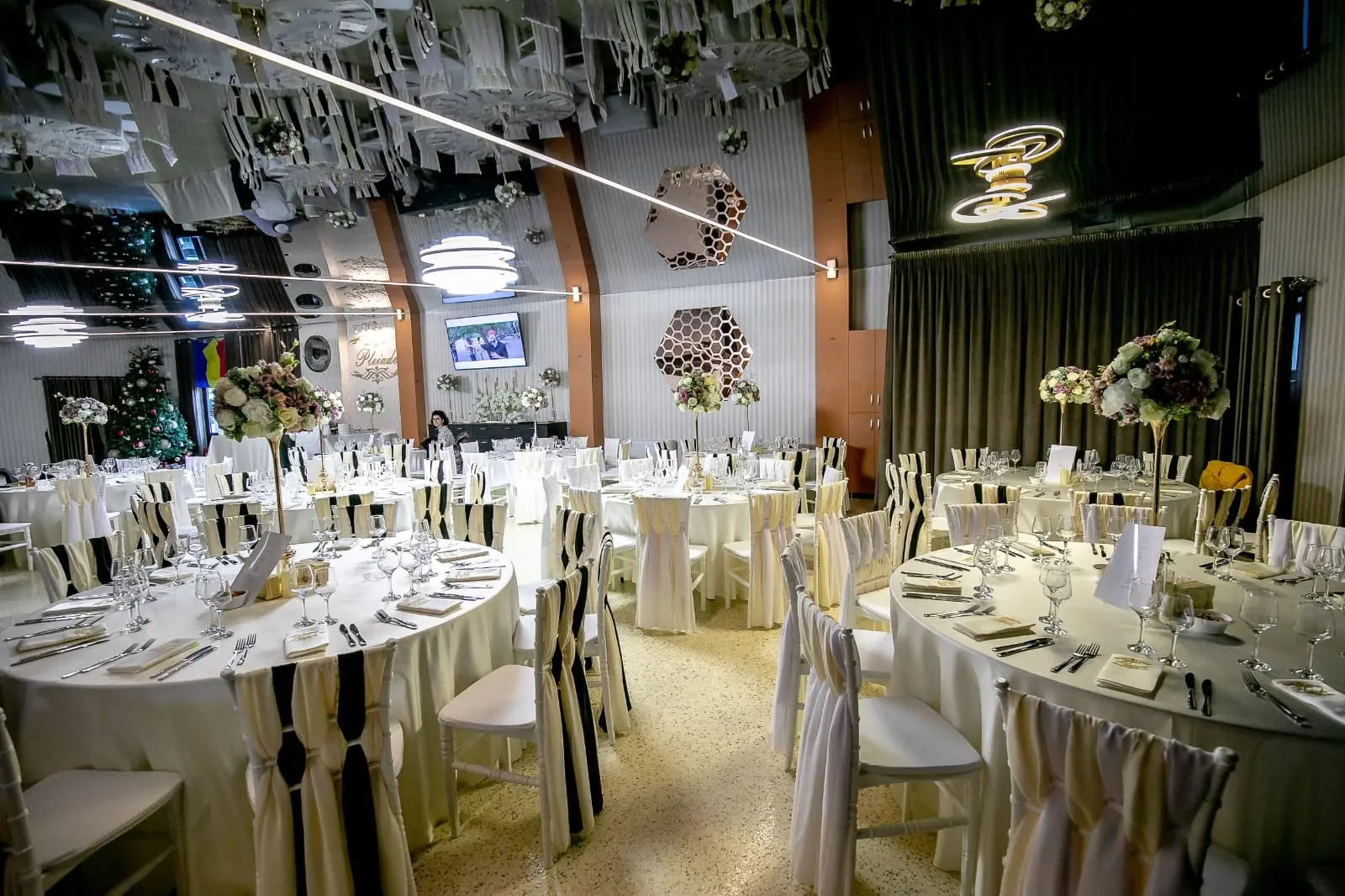 Sala aranjată cu mese rotunde și lustre LED. În fundal, un brad de Crăciun.