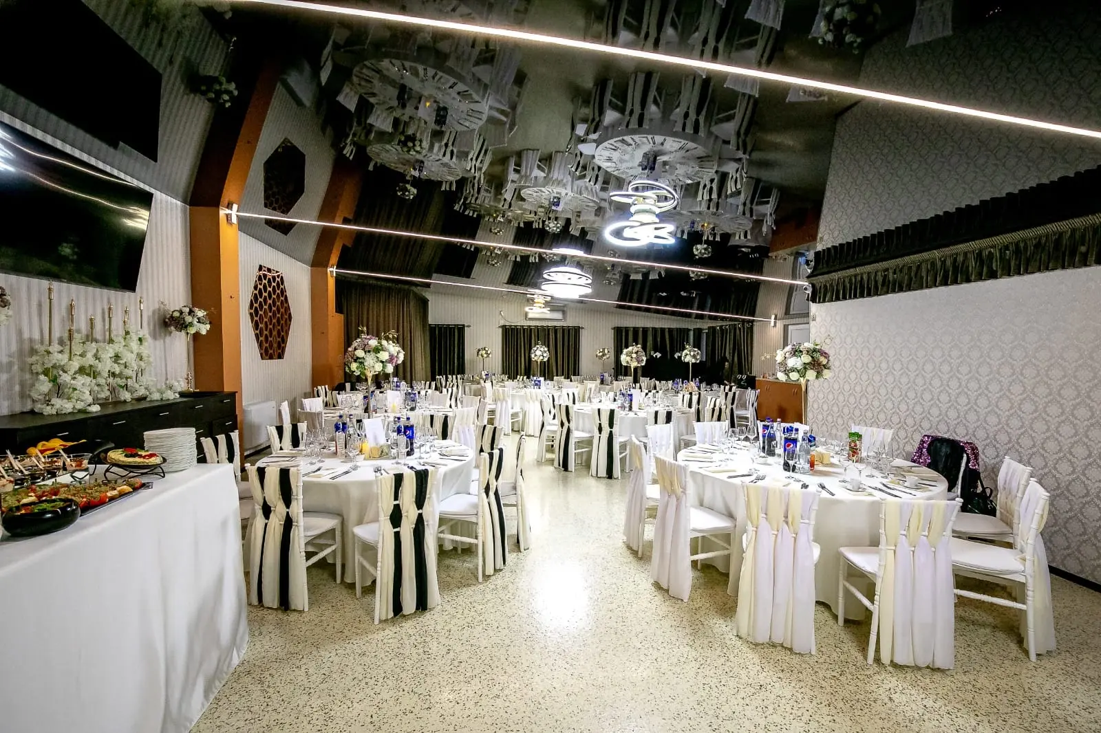 Sala cu mese rotunde îmbrăcate în alb și scaune albe cu eșarfe crem și negre. În stânga, o masă cu aperitive, iar deasupra, tavanul negru lucios cu linii LED și lustre circulare aprinse.