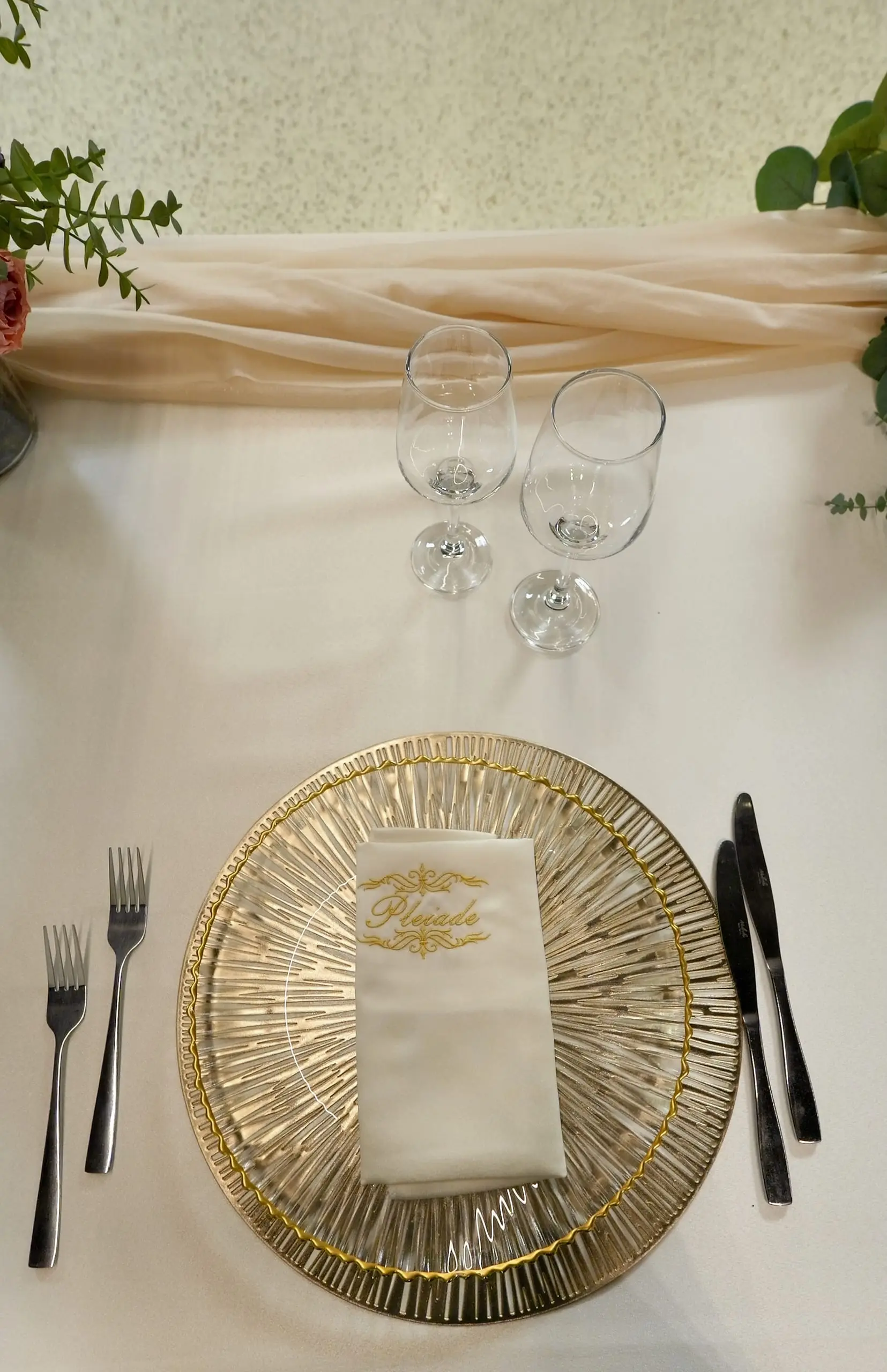 Tacâmul unui invitat, văzut de sus: farfurie de prezentare aurie, șervet crem cu numele Pleiade brodat auriu, două pahare, două furculițe și două cuțite.